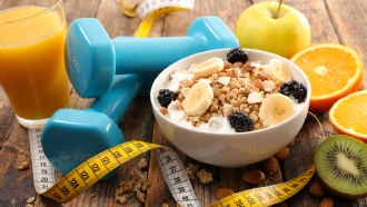 Taça de cereais com iogurte, em cima de uma mesa, com maçãs e laranja cortada ao lado, um copo com sumo de laranja, uma fita métrica e dois pesos para os braços.